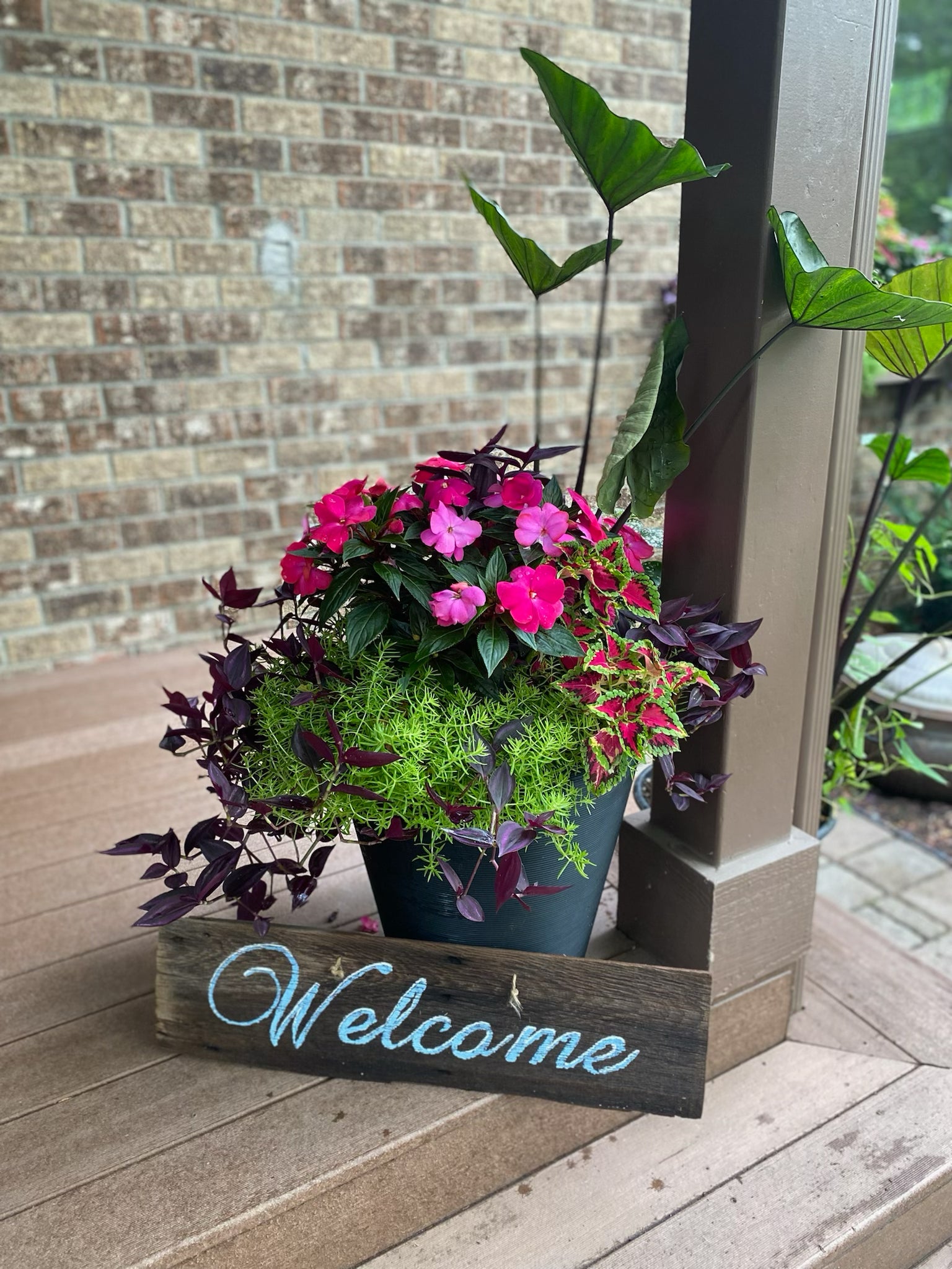 Potted plant with pink flowers on a wooden 'Welcome' sign in front of a brick wall.