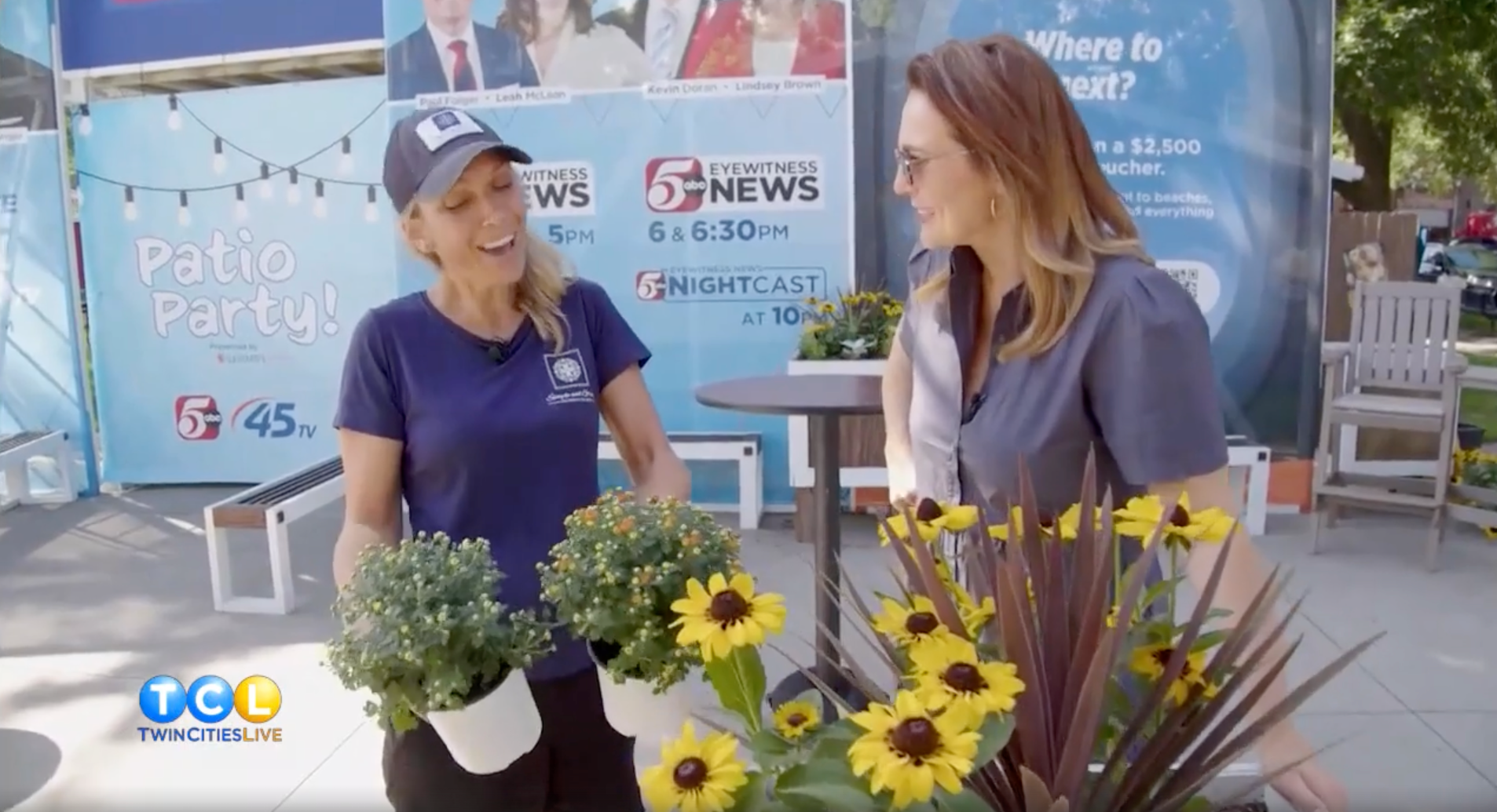 Two women standing next to potted plants with a news station backdrop