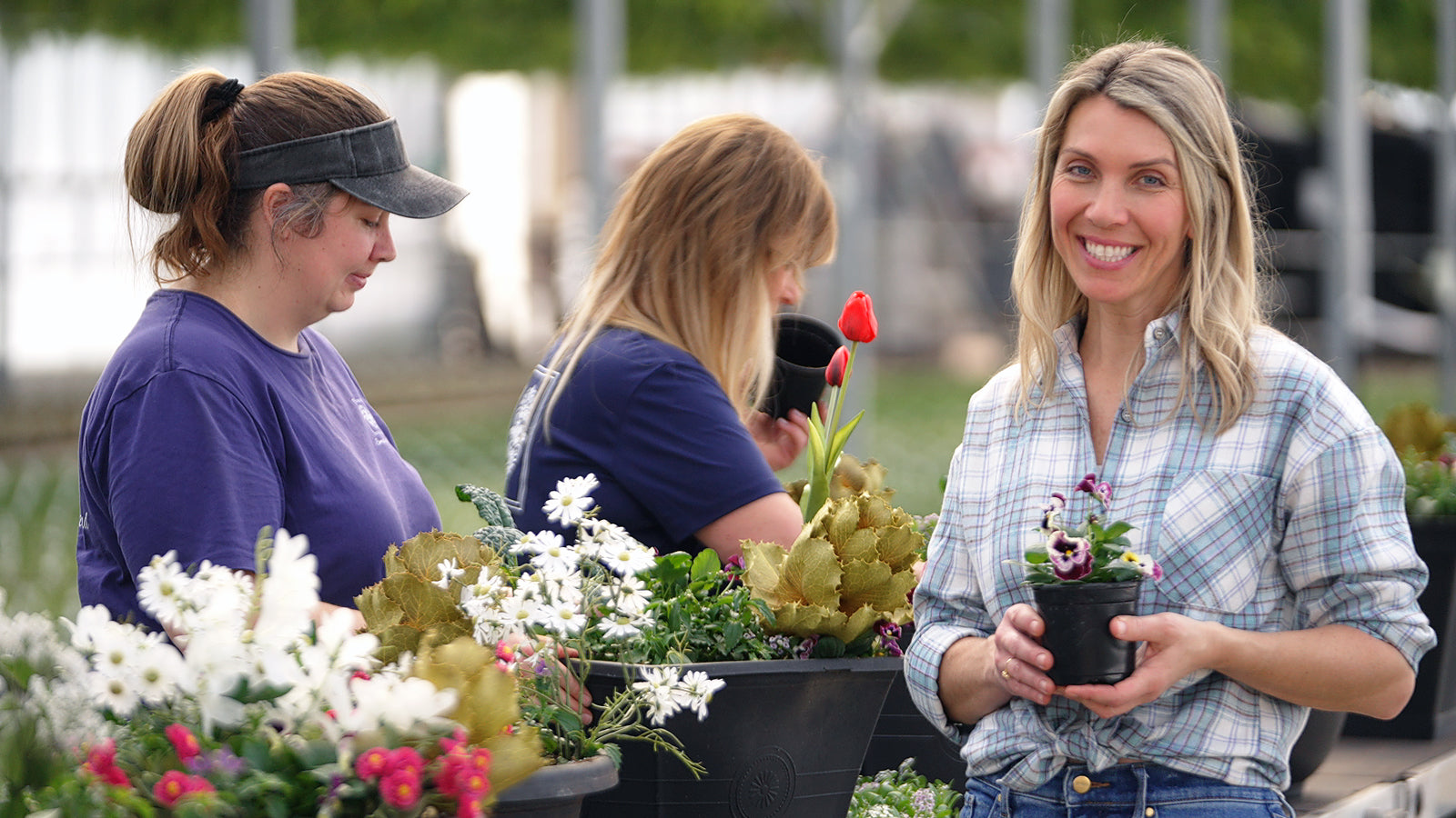 Three women at a garden center, with one holding a potted plant.
