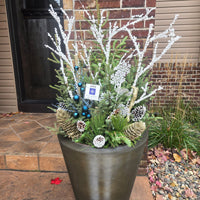 A large dark gray concrete planter filled with holiday greenery, including white berry branches, pinecones, blue berries, and small decorative tags, placed outdoors on a brick patio in front of a house.